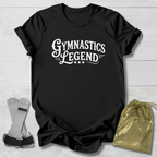Black t-shirt with 'Gymnastics Legend' text on a light wooden surface with socks and a drawstring bag.