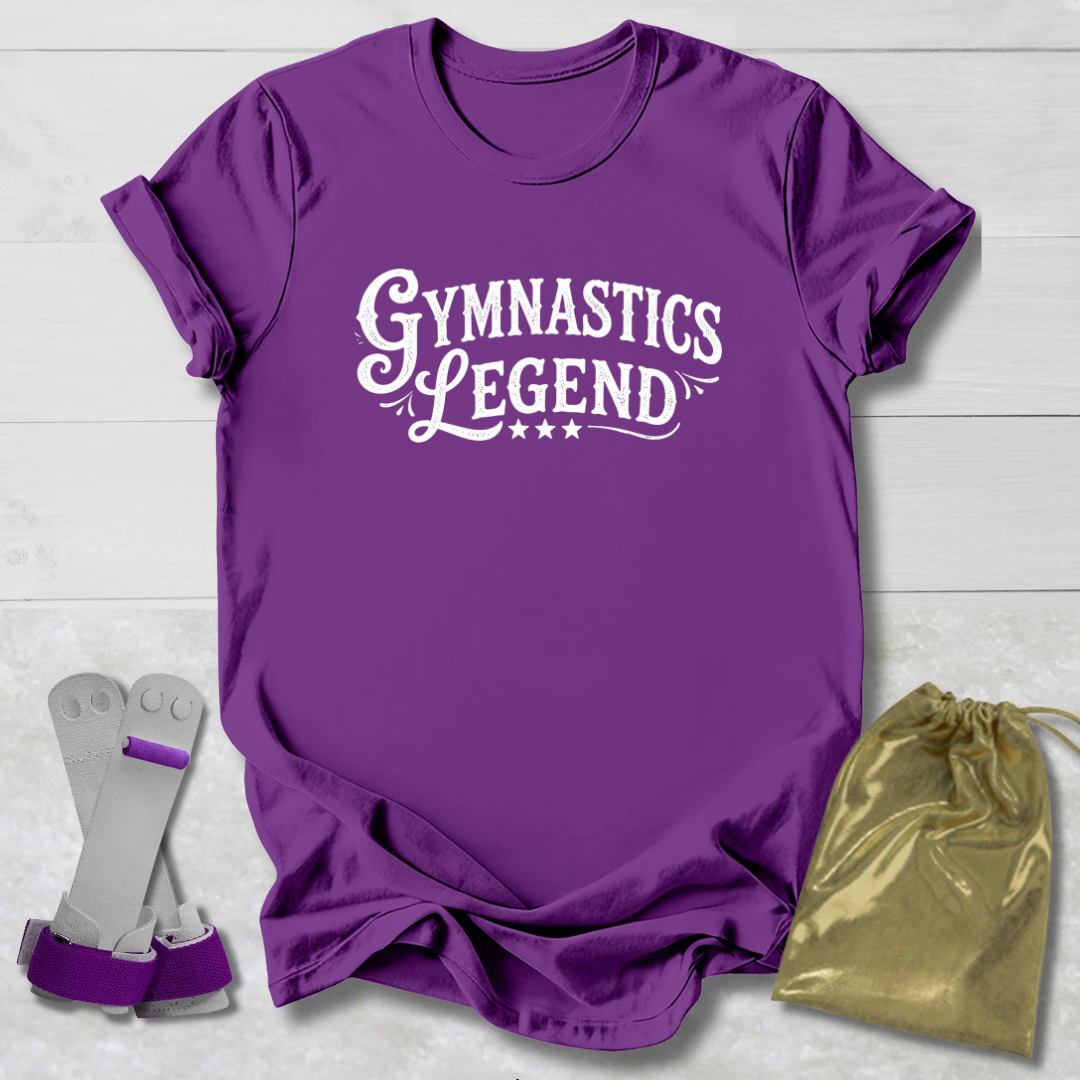 Purple t-shirt with 'Gymnastics Legend' text, gray socks, and gold drawstring bag on a light wooden surface.