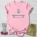 Pink t-shirt with a graphic of a gymnast on a balance beam, accompanied by socks and a drawstring bag on a light wooden surface.