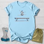 Light blue t-shirt with a graphic of a gymnast on a balance beam, paired with socks and a drawstring bag on a wooden surface.
