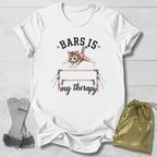 White t-shirt with a graphic of a cat on bars and text, placed on a light wooden surface with socks and a drawstring bag.
