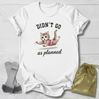 White t-shirt with a cat graphic and text 'Didn't go as planned' on a light wooden surface, with socks and a drawstring bag.