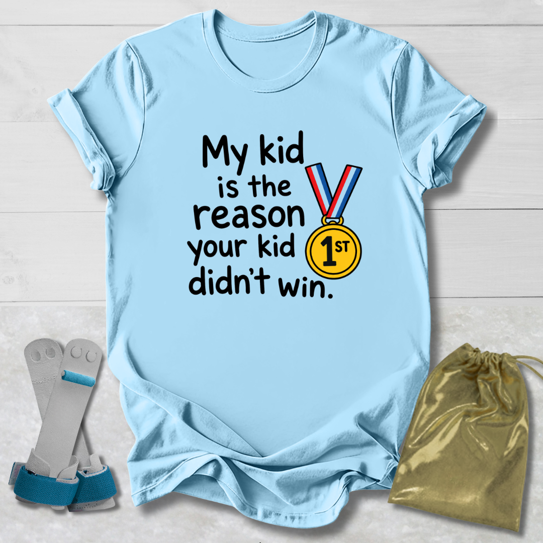 A light blue graphic T-shirt laid flat on a light wood background, with the text 'My kid is the reason your kid didn't win.' and a medal illustration.