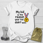 A white short-sleeve graphic T-shirt laid flat on a light wood background, with black text reading 'My kid is the reason your kid didn't win.' and a medal graphic featuring red, white, and blue ribbon bars and a yellow '1st' medallion.