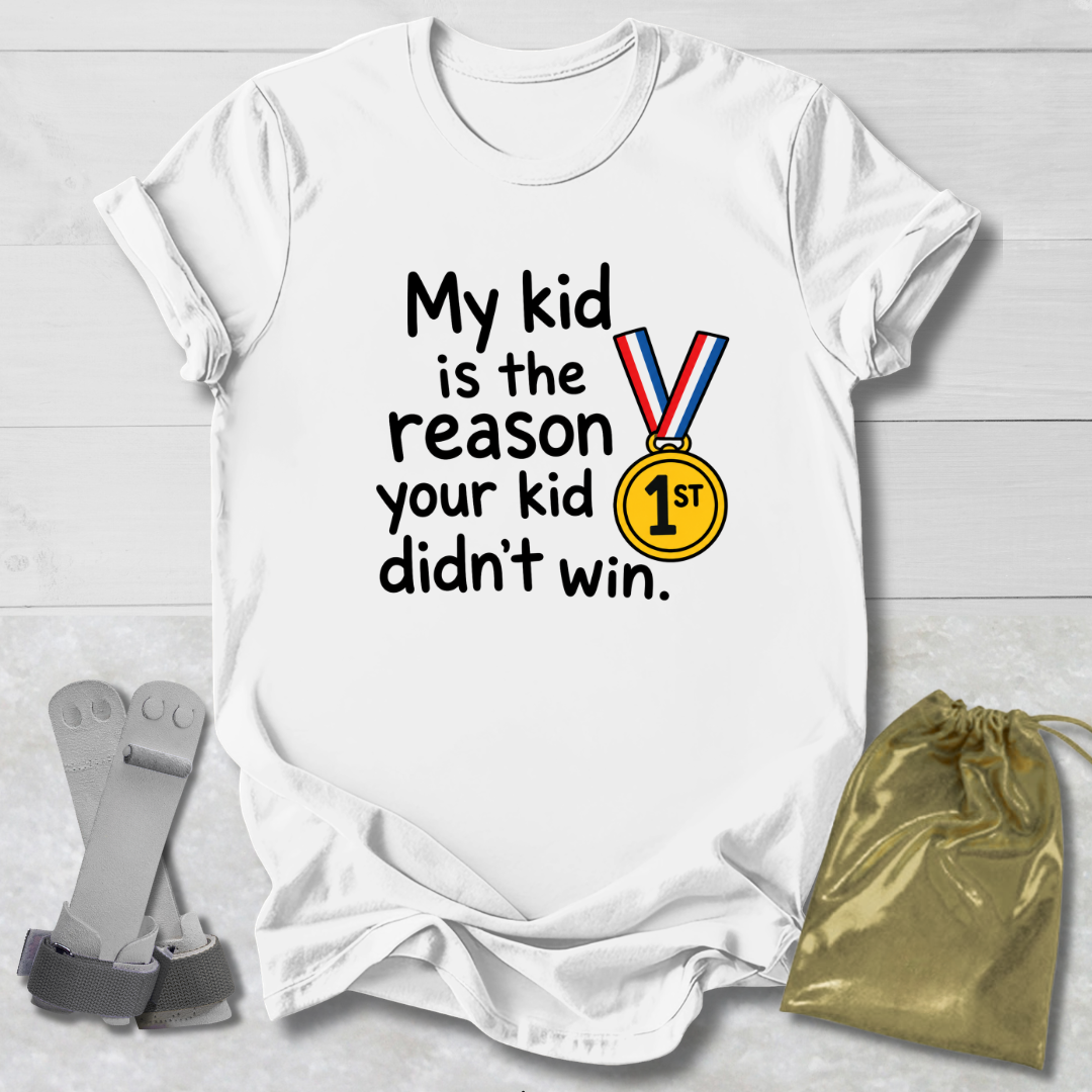 A white short-sleeve graphic T-shirt laid flat on a light wood background, with black text reading 'My kid is the reason your kid didn't win.' and a medal graphic featuring red, white, and blue ribbon bars and a yellow '1st' medallion.