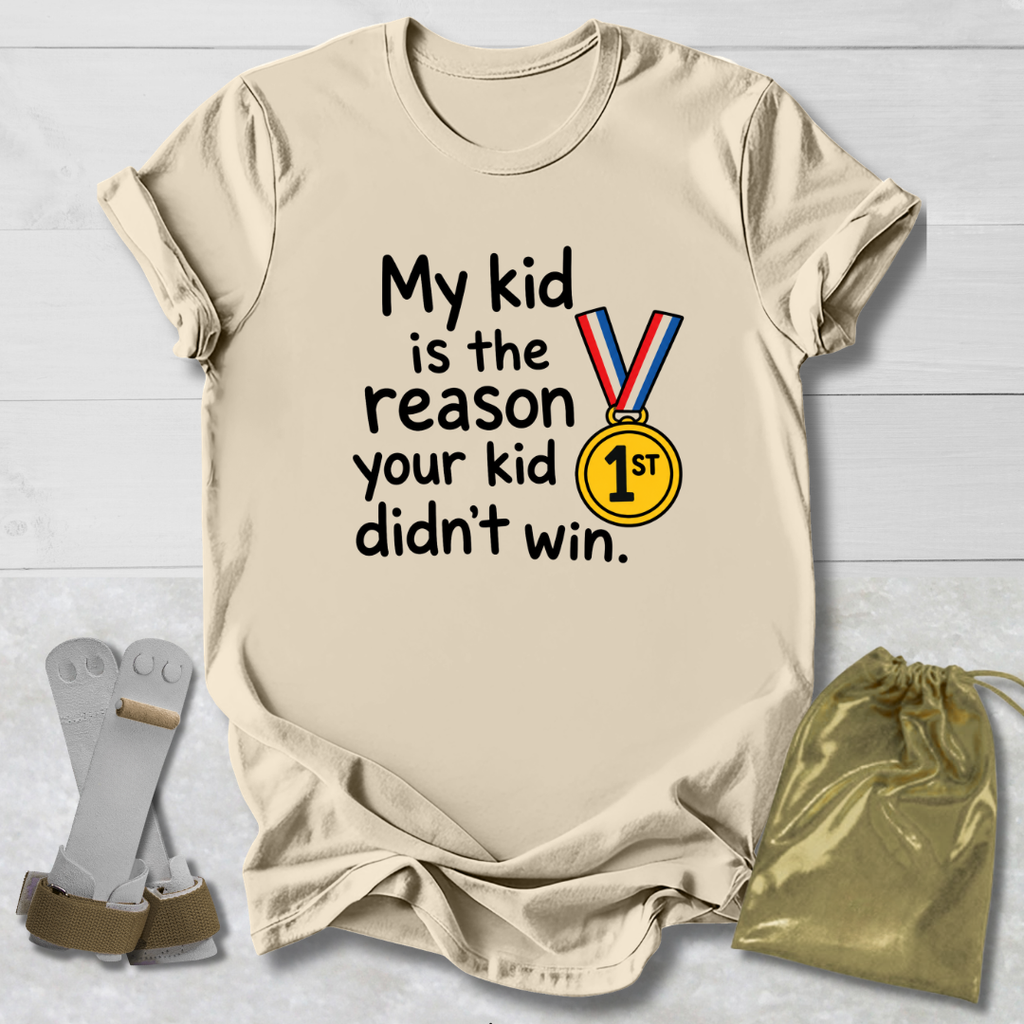 A beige graphic t-shirt laid flat on a white wood background, featuring the text 'My kid is the reason your kid didn't win.' alongside a medal graphic.