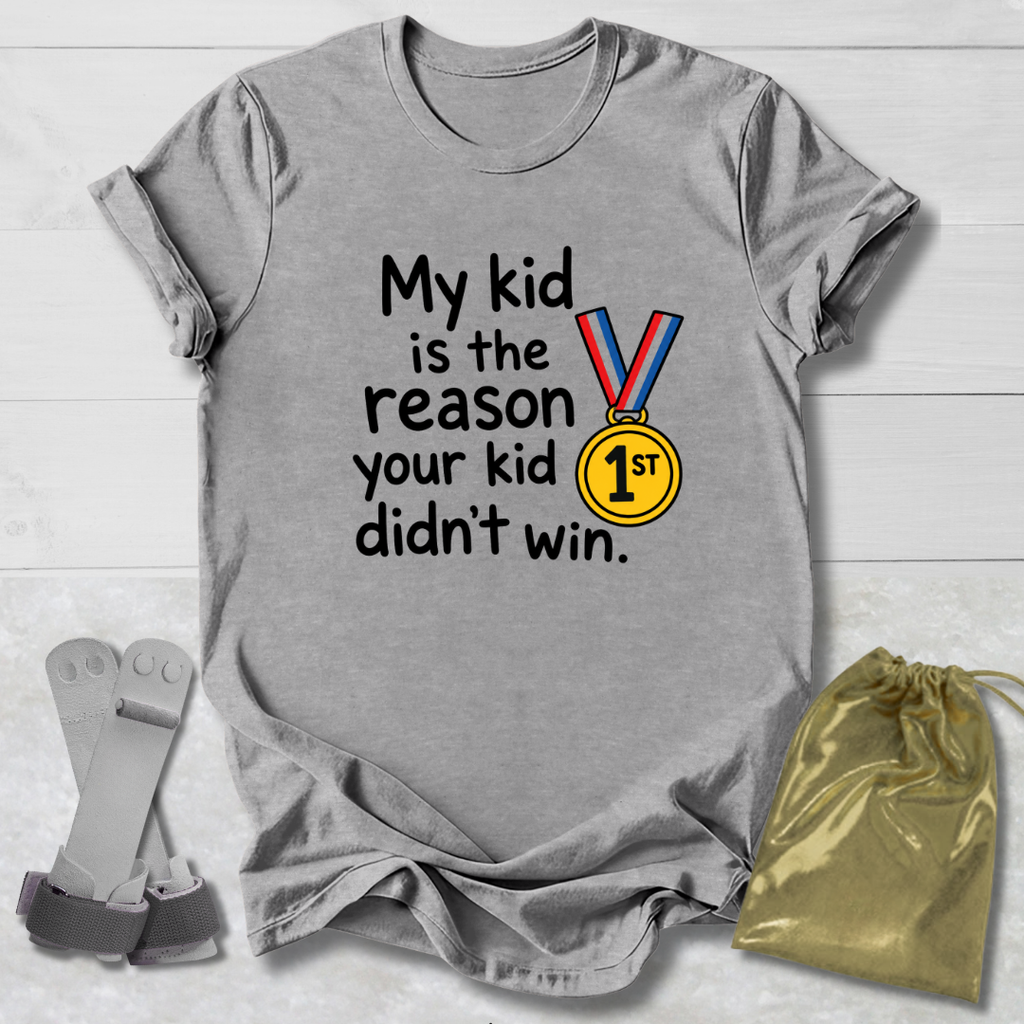 A heather gray graphic T-shirt laid flat on a white wood background, printed with the phrase 'My kid is the reason your kid didn't win.' in black text beside a medal graphic with red, blue, and yellow ribbon and a gold '1st' medallion.