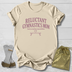 A beige graphic T-shirt laid flat on a light wood background, printed with the phrase 'RELUCTANT GYMNASTICS MOM' and a small bench graphic in dusty pink. A pair of gymnastics straps and a shiny gold drawstring bag are placed beside the shirt as styling props.