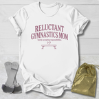 A white short-sleeve T-shirt laid flat on a light wood background, printed with the phrase 'RELUCTANT GYMNASTICS MOM' and a small bench graphic in dusty pink. A gray gymnastics strap and a shiny gold drawstring pouch are placed beside the shirt as styling props.