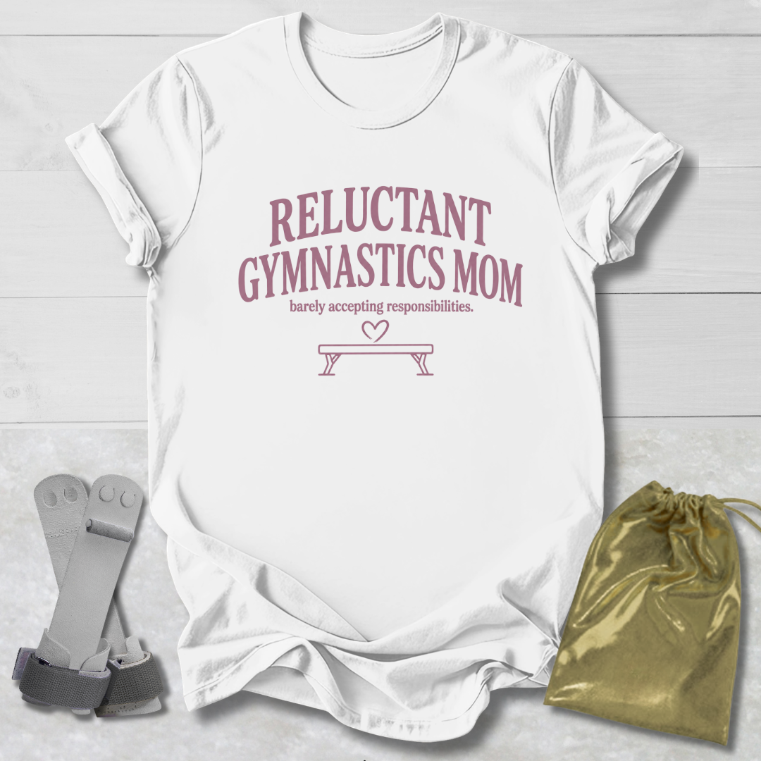 A white short-sleeve T-shirt laid flat on a light wood background, printed with the phrase 'RELUCTANT GYMNASTICS MOM' and a small bench graphic in dusty pink. A gray gymnastics strap and a shiny gold drawstring pouch are placed beside the shirt as styling props.