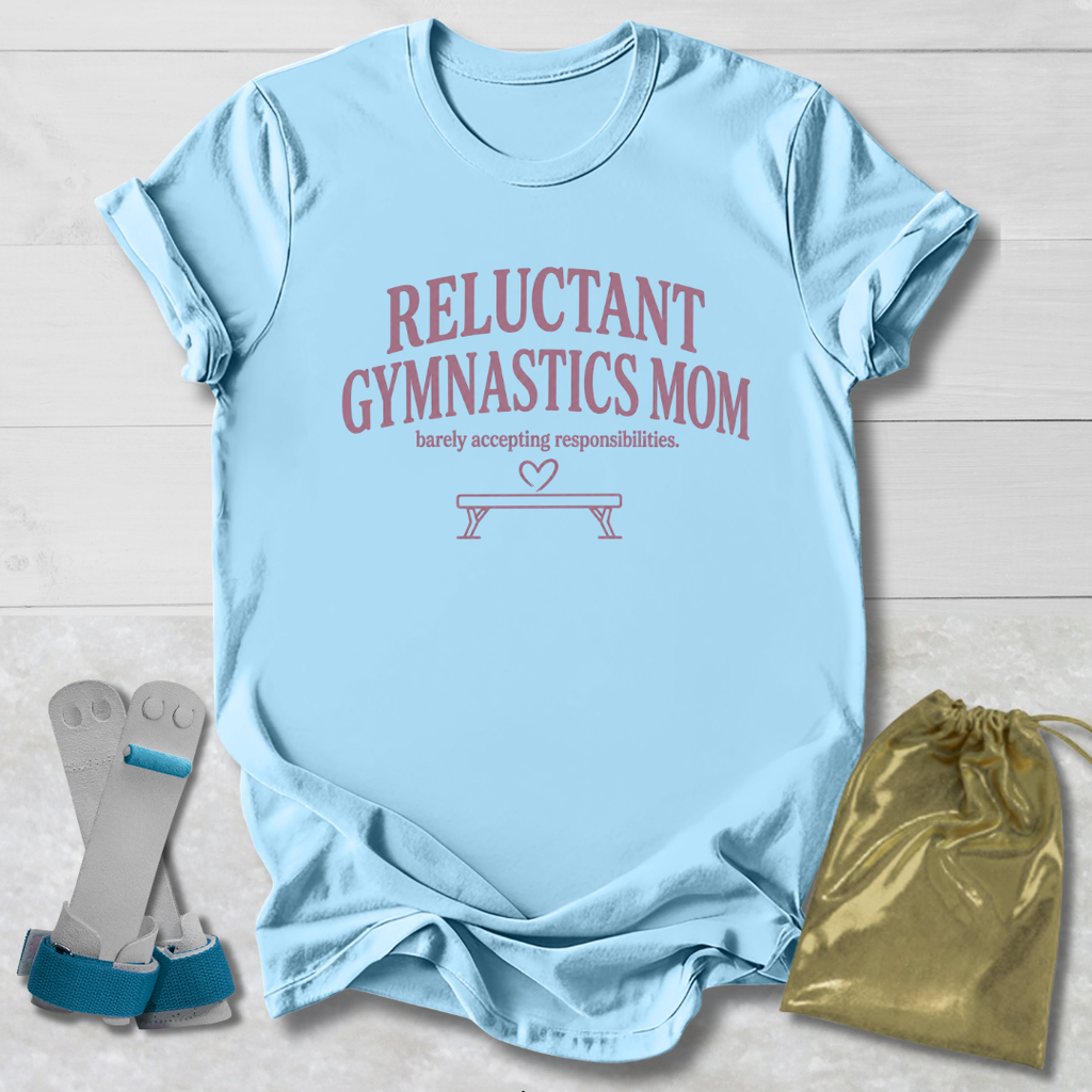 A light blue graphic t-shirt laid flat on a white wood background, featuring the text 'RELUCTANT GYMNASTICS MOM' in dusty pink. Accompanying props include a pair of gymnastics bars with blue straps and a shiny gold drawstring bag.