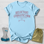 A light blue graphic t-shirt laid flat on a white wood background, featuring the text 'RELUCTANT GYMNASTICS MOM' in dusty pink. Accompanying props include a pair of gymnastics bars with blue straps and a shiny gold drawstring bag.