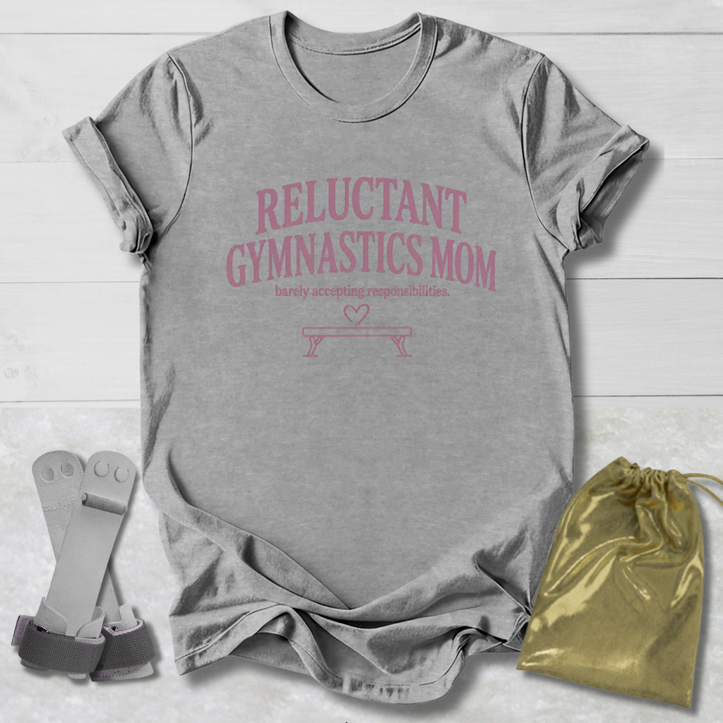 A heather gray short-sleeve t-shirt with a red crown graphic and the words 'GYMNASTICS Queen' printed on the front, shown in a flat-lay arrangement with a pair of gray wrist straps and a gold drawstring pouch on a white wood background.