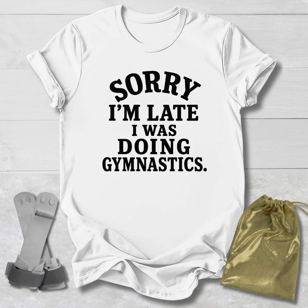 A white short-sleeve graphic T-shirt laid flat on a light wood background, printed with the phrase 'SORRY I'M LATE I WAS DOING GYMNASTICS.' in bold black text.