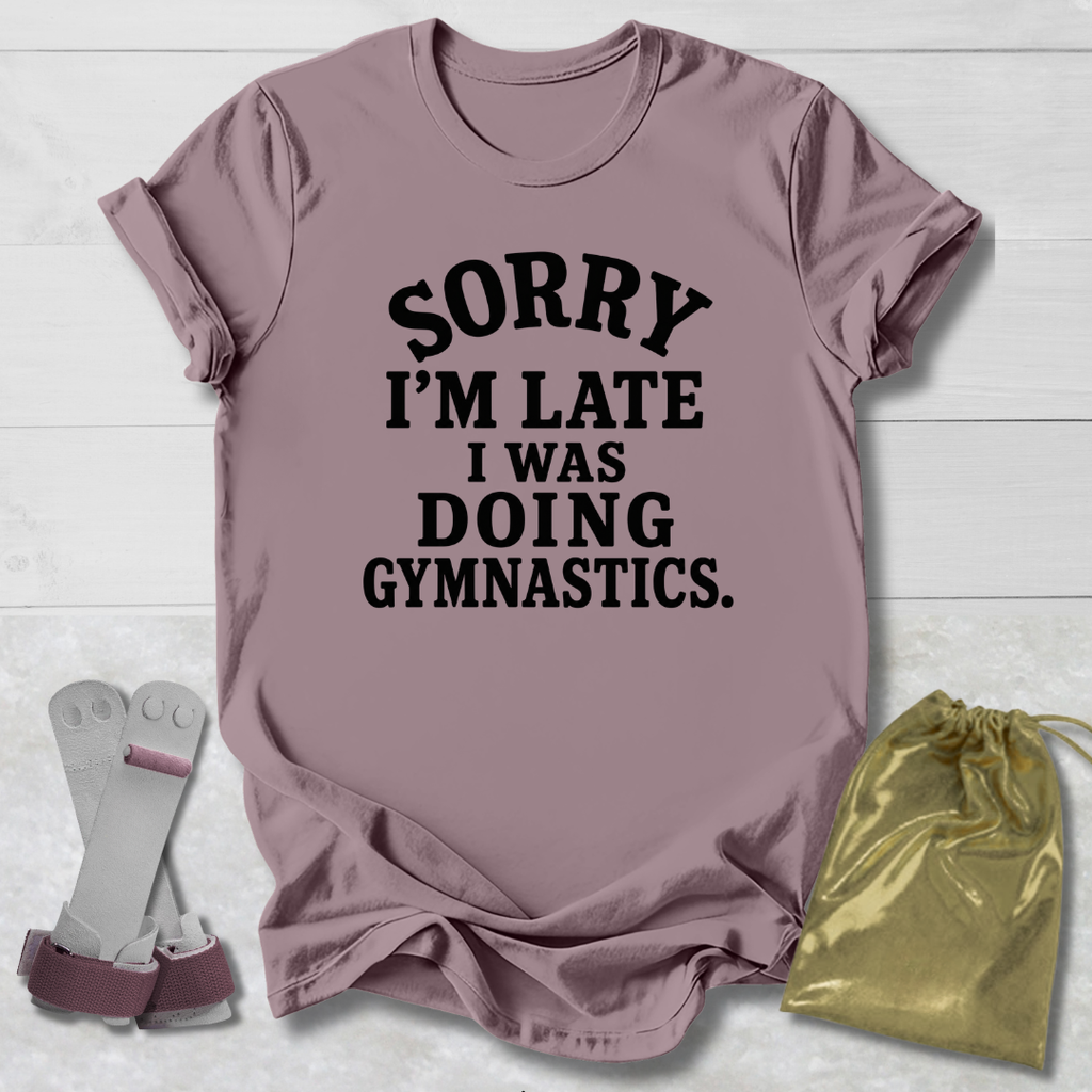 A mauve short-sleeve t-shirt with black text reading 'SORRY I'M LATE I WAS DOING GYMNASTICS.' laid flat on a white wood background, styled with a pair of gymnastics bars and a gold drawstring bag.