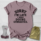 A mauve short-sleeve t-shirt with black text reading 'SORRY I'M LATE I WAS DOING GYMNASTICS.' laid flat on a white wood background, styled with a pair of gymnastics bars and a gold drawstring bag.