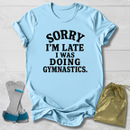 A light blue short-sleeve t-shirt with black text reading 'SORRY I'M LATE I WAS DOING GYMNASTICS.' laid flat on a white wood background, styled with gymnastics straps and a gold drawstring bag.