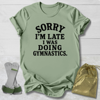 A sage green graphic t-shirt with black text reading 'SORRY I'M LATE I WAS DOING GYMNASTICS.' laid flat on a white wood background, styled with a pair of gymnastics bars and a gold drawstring bag.