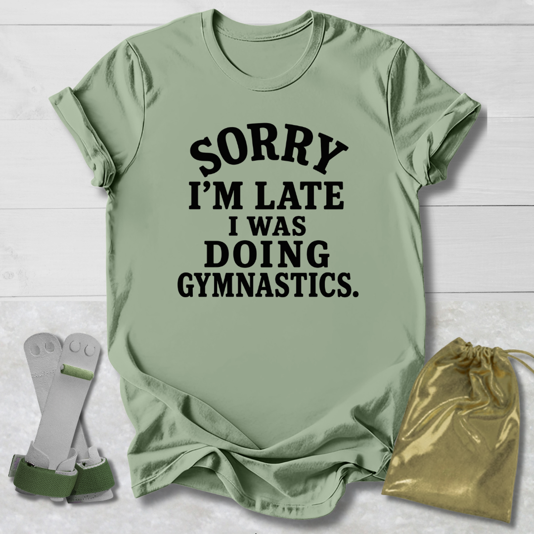 A sage green graphic t-shirt with black text reading 'SORRY I'M LATE I WAS DOING GYMNASTICS.' laid flat on a white wood background, styled with a pair of gymnastics bars and a gold drawstring bag.