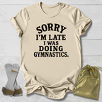 A beige graphic t-shirt with the text 'SORRY I'M LATE I WAS DOING GYMNASTICS.' printed in black.