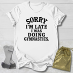 A white short-sleeve graphic T-shirt laid flat on a light wood background, printed with the phrase 'SORRY I'M LATE I WAS DOING GYMNASTICS.' in bold black text.