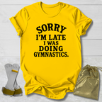 A bright yellow short-sleeve t-shirt with black text reading 'SORRY I'M LATE I WAS DOING GYMNASTICS.' laid flat on a white wood background, styled with a pair of gymnastics bars and a metallic gold drawstring bag.