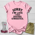 A light pink short-sleeve t-shirt with bold black text reading 'SORRY I'M LATE I WAS DOING GYMNASTICS.' laid flat on a white wood background, styled with gymnastics accessories including a gold drawstring bag and a pair of pink-and-white hand grips.