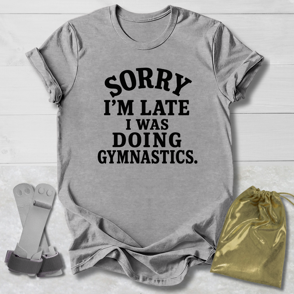 A heather gray short-sleeve t-shirt with black text reading 'SORRY I'M LATE I WAS DOING GYMNASTICS.' laid flat on a white wood background, styled with a pair of gymnastics bars and a small gold drawstring bag.