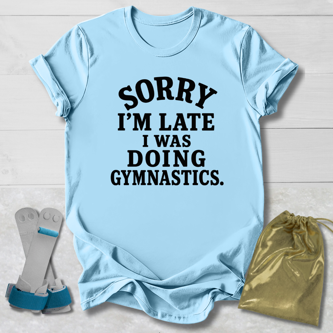 A light blue short-sleeve t-shirt with black text reading 'SORRY I'M LATE I WAS DOING GYMNASTICS.' laid flat on a white wood background, styled with gymnastics straps and a gold drawstring bag.