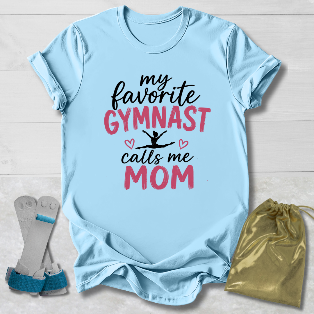 A light blue graphic T-shirt laid flat on a white wood background, with the text 'my favorite GYMNAST calls me MOM' in black and pink lettering. A gymnast silhouette and small hearts appear in the design.