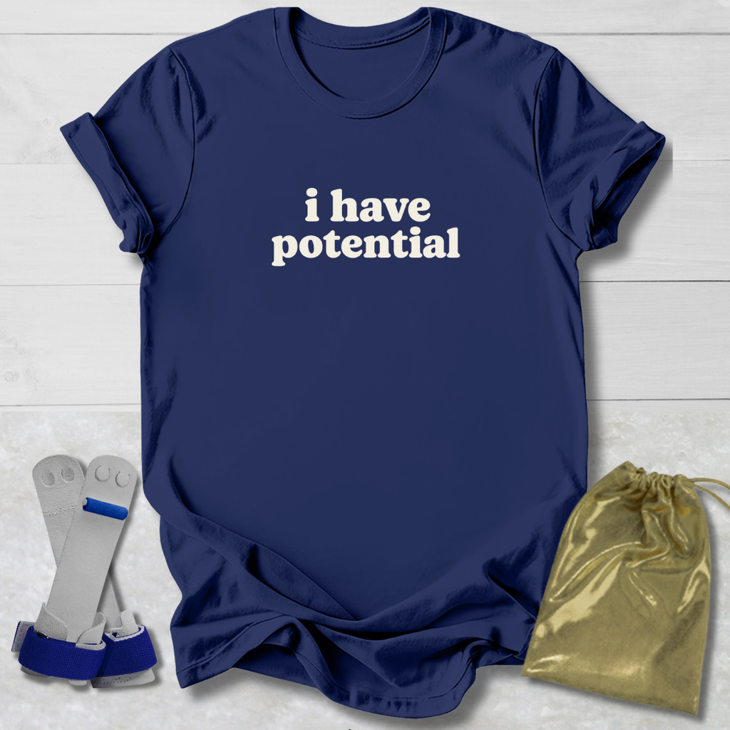 A navy blue short-sleeve T-shirt with the phrase 'i have potential' printed in white across the chest, laid flat on a light wood background. A pair of gray and blue wrist/ankle cuffs and a shiny gold drawstring pouch are placed beside the shirt as included accessories or styling props.