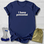 A navy blue short-sleeve T-shirt with the phrase 'i have potential' printed in white across the chest, laid flat on a light wood background. A pair of gray and blue wrist/ankle cuffs and a shiny gold drawstring pouch are placed beside the shirt as included accessories or styling props.