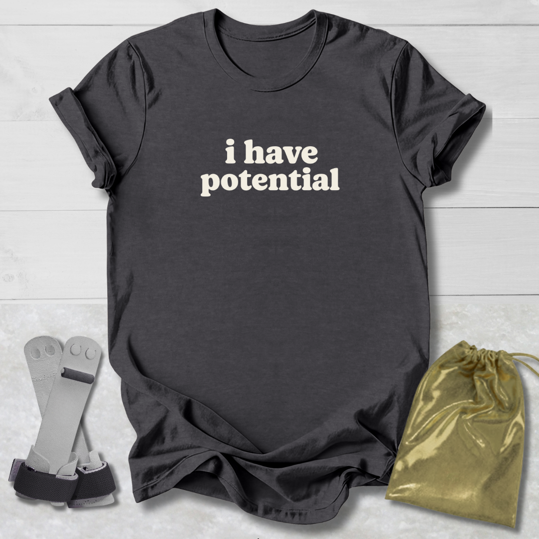 A dark charcoal gray t-shirt with the phrase 'i have potential' printed in white text across the chest, styled in a flat-lay arrangement with a pair of gray arm straps and a shiny gold drawstring pouch on a light gray wood plank background.