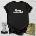 A black short-sleeve T-shirt with the phrase 'i have potential' printed in white across the chest, laid flat on a light wood background. A pair of gray and black wrist wraps and a small shiny gold drawstring pouch are placed beside the shirt.