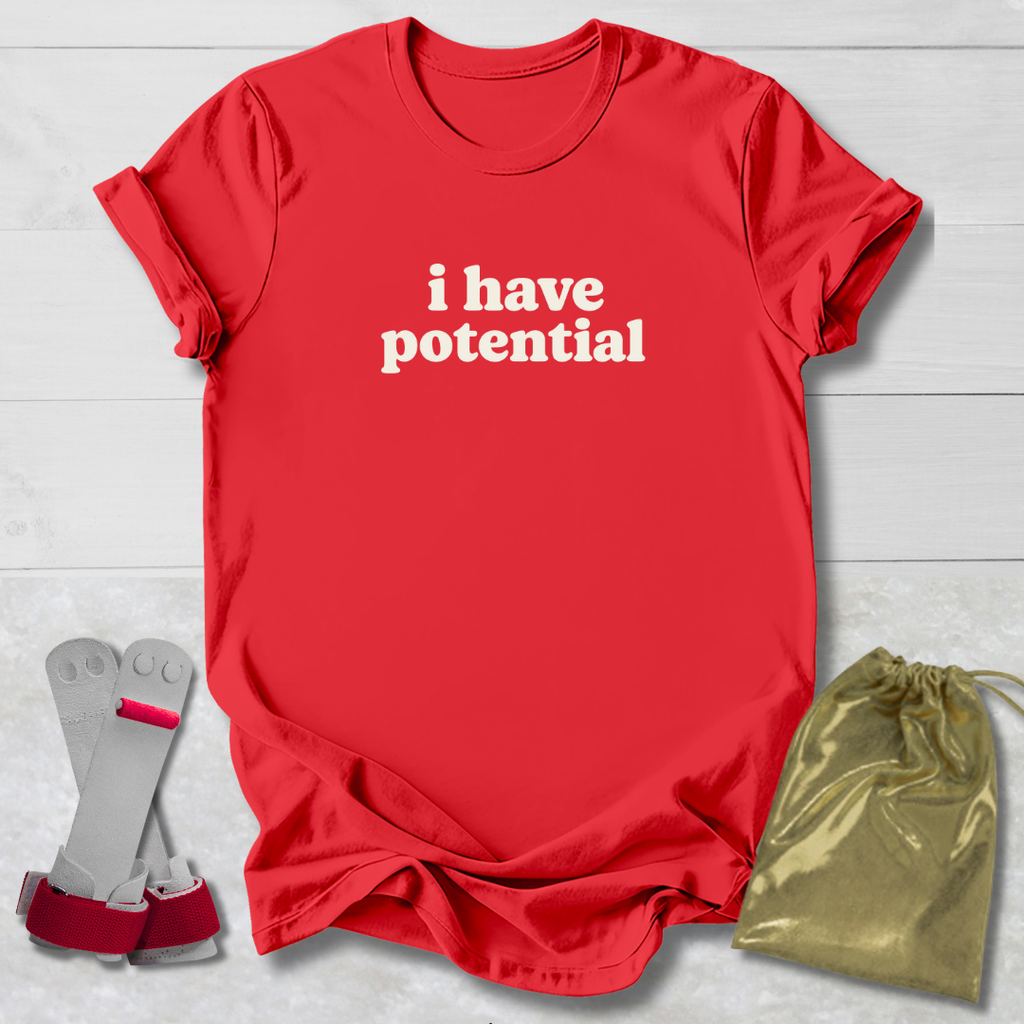A red short-sleeve T-shirt with the phrase 'i have potential' printed in white lowercase letters across the chest, laid flat on a light wood background. A pair of gray and red arm cuffs and a shiny gold drawstring pouch are placed beside the shirt as included accessories or packaging.