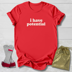 A red short-sleeve T-shirt with the phrase 'i have potential' printed in white lowercase letters across the chest, laid flat on a light wood background. A pair of gray and red arm cuffs and a shiny gold drawstring pouch are placed beside the shirt as included accessories or packaging.