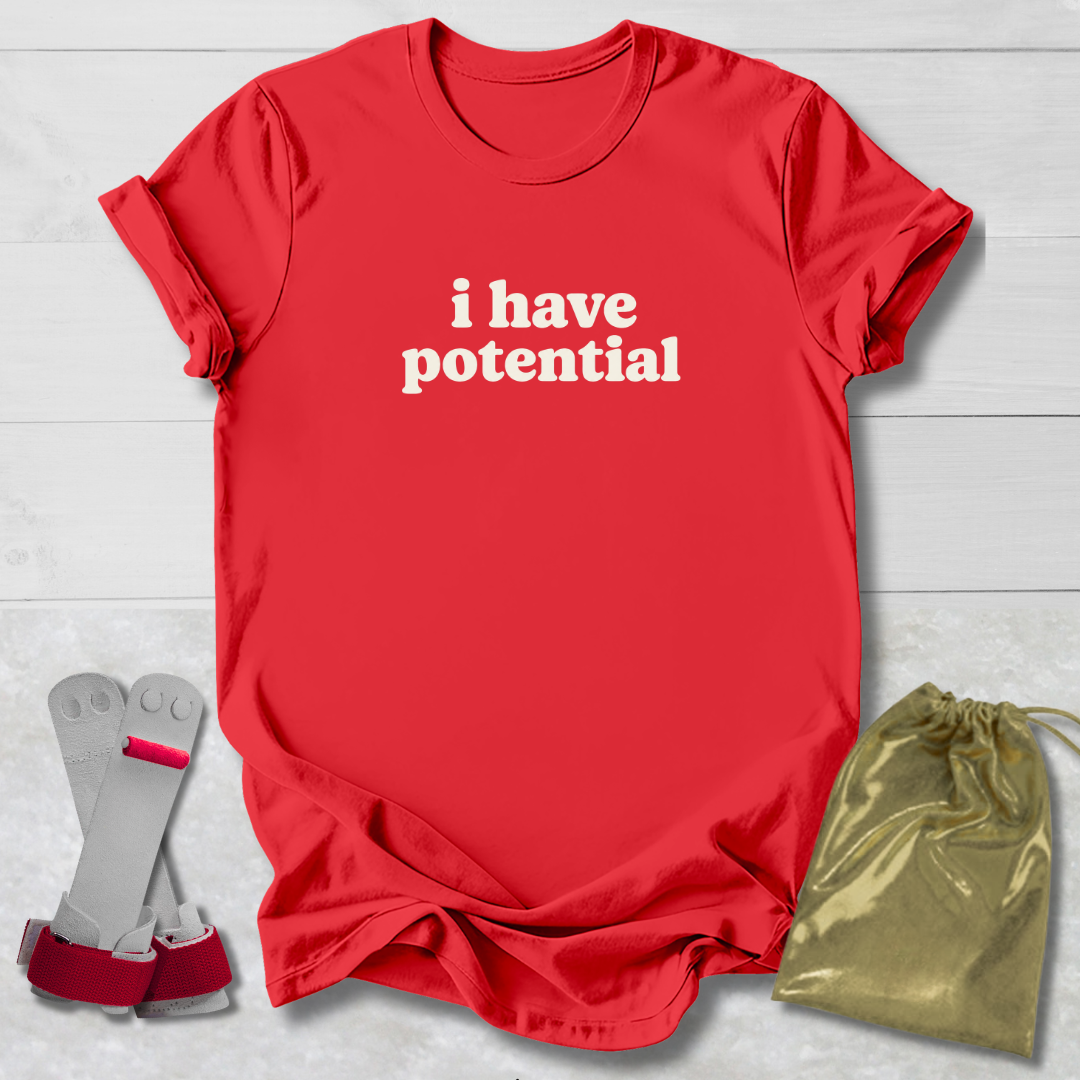A red short-sleeve T-shirt with the phrase 'i have potential' printed in white lowercase letters across the chest, laid flat on a light wood background. A pair of gray and red arm cuffs and a shiny gold drawstring pouch are placed beside the shirt as included accessories or packaging.