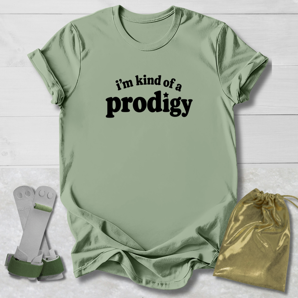 A sage green graphic t-shirt with the text 'i'm kind of a prodigy' printed in black on the front, styled in a flat-lay with a gold drawstring pouch and white cleats on a white wood background.