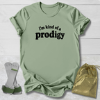 A sage green graphic t-shirt with the text 'i'm kind of a prodigy' printed in black on the front, styled in a flat-lay with a gold drawstring pouch and white cleats on a white wood background.