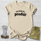 A beige graphic t-shirt with the text 'i'm kind of a prodigy' printed in black on the front, styled in a flat-lay with a gold satin drawstring bag and white-framed glasses on a white wood background.