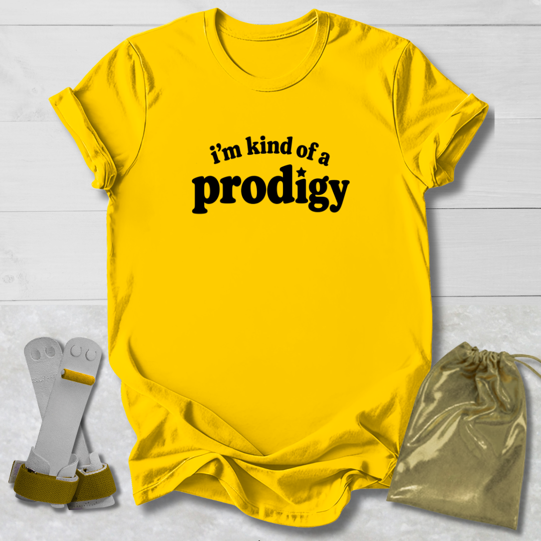 A bright yellow graphic t-shirt with the black text 'i'm kind of a prodigy' printed on the chest, styled in a flat-lay with a gold drawstring pouch and a pair of white and gold glasses on a white wood background.