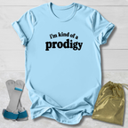 A light blue graphic t-shirt with the text 'i'm kind of a prodigy' printed in black on the front, styled in a flat-lay with a gold drawstring bag and a pair of gray and blue support braces on a white wood background.