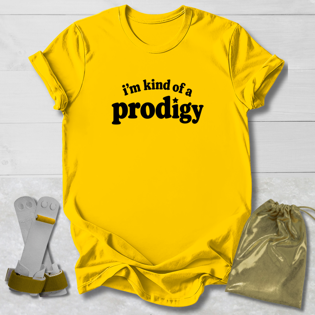 A bright yellow graphic t-shirt with the black text 'i'm kind of a prodigy' printed on the chest, styled in a flat-lay with a gold drawstring pouch and a pair of white and gold glasses on a white wood background.