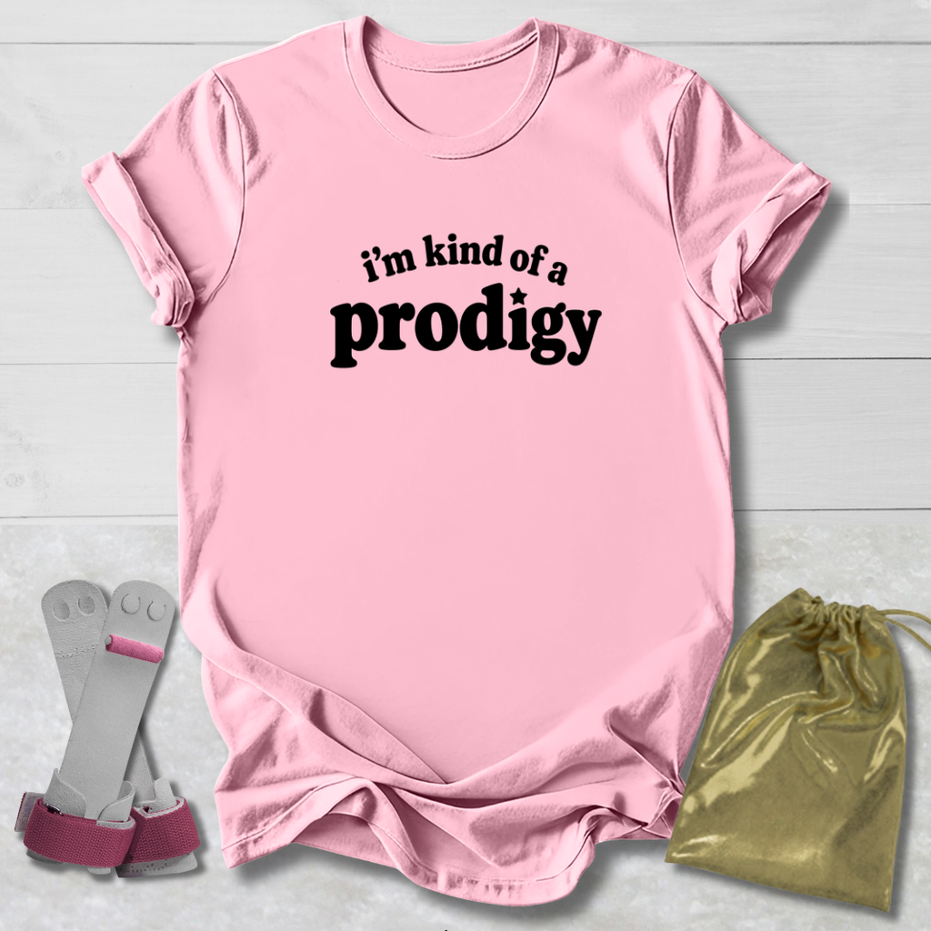 A light pink short-sleeve t-shirt with the black text 'i'm kind of a prodigy' printed on the chest, styled in a flat-lay with a gold drawstring bag and gray-and-pink socks on a white wood background.