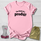 A light pink short-sleeve t-shirt with the black text 'i'm kind of a prodigy' printed on the chest, styled in a flat-lay with a gold drawstring bag and gray-and-pink socks on a white wood background.