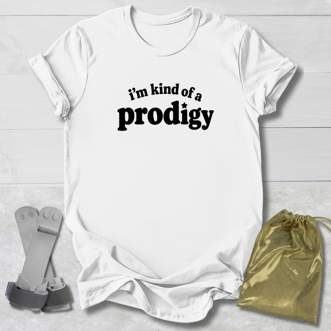 A white short-sleeve T-shirt laid flat on a light wood background, printed with the black text 'i'm kind of a prodigy.' Two small props are placed beside it: a gray strap-like accessory on the left and a shiny gold drawstring pouch on the right.