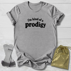 A heather gray short-sleeve t-shirt with the black printed text 'i'm kind of a prodigy' on the chest, laid flat on a white wood background alongside a gold drawstring bag and a pair of gray-and-white sandals.