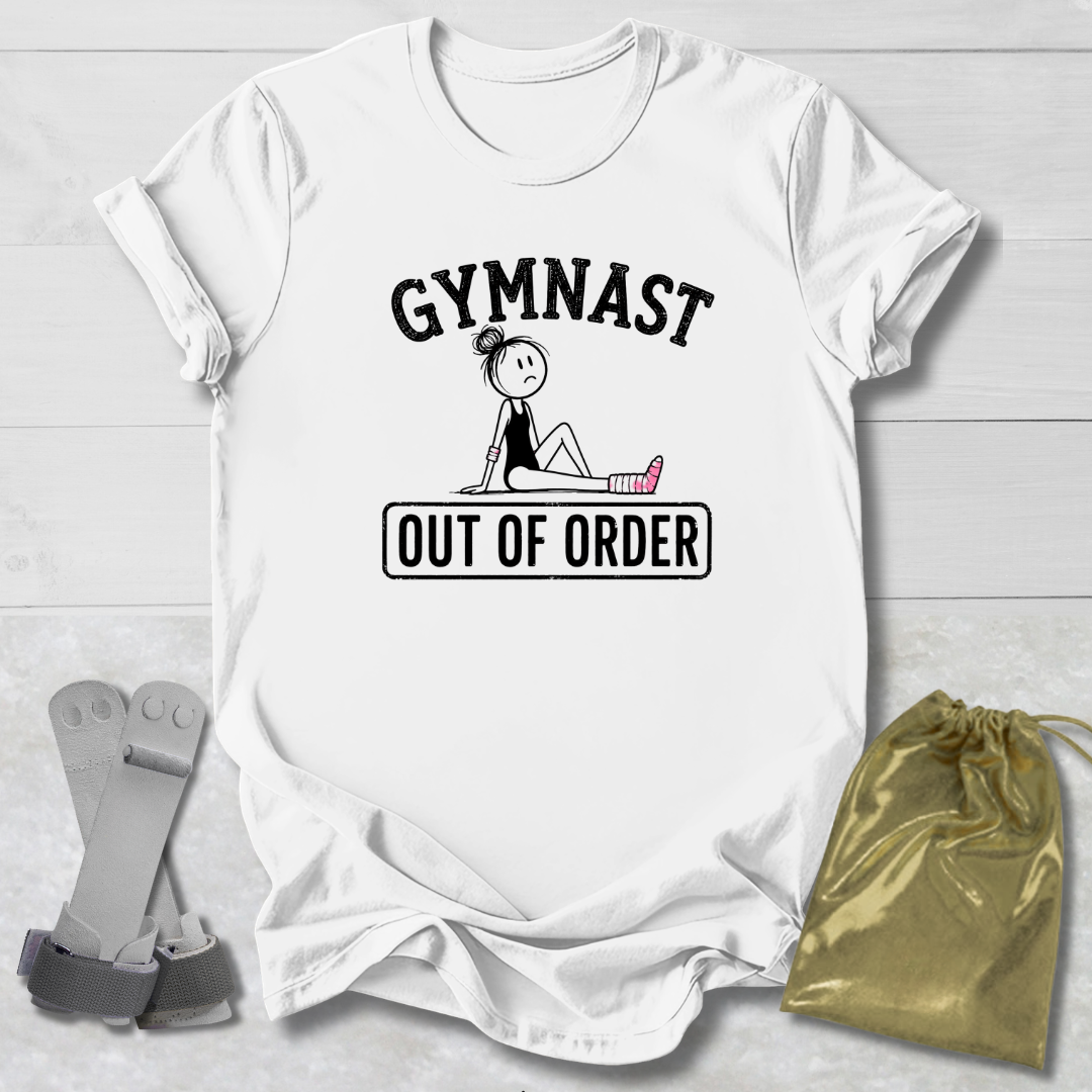 A white short-sleeve graphic T-shirt laid flat on a light wood background, featuring the words 'GYMNAST OUT OF ORDER' with a simple line drawing of a gymnast. A pair of gray straps and a gold drawstring bag are placed beside the shirt as styling props.