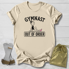 A beige graphic t-shirt laid flat on a white wood background, featuring the text 'GYMNAST OUT OF ORDER' with a simple gymnast illustration. A pair of gymnast straps and a gold drawstring bag are placed beside the shirt as styling props.