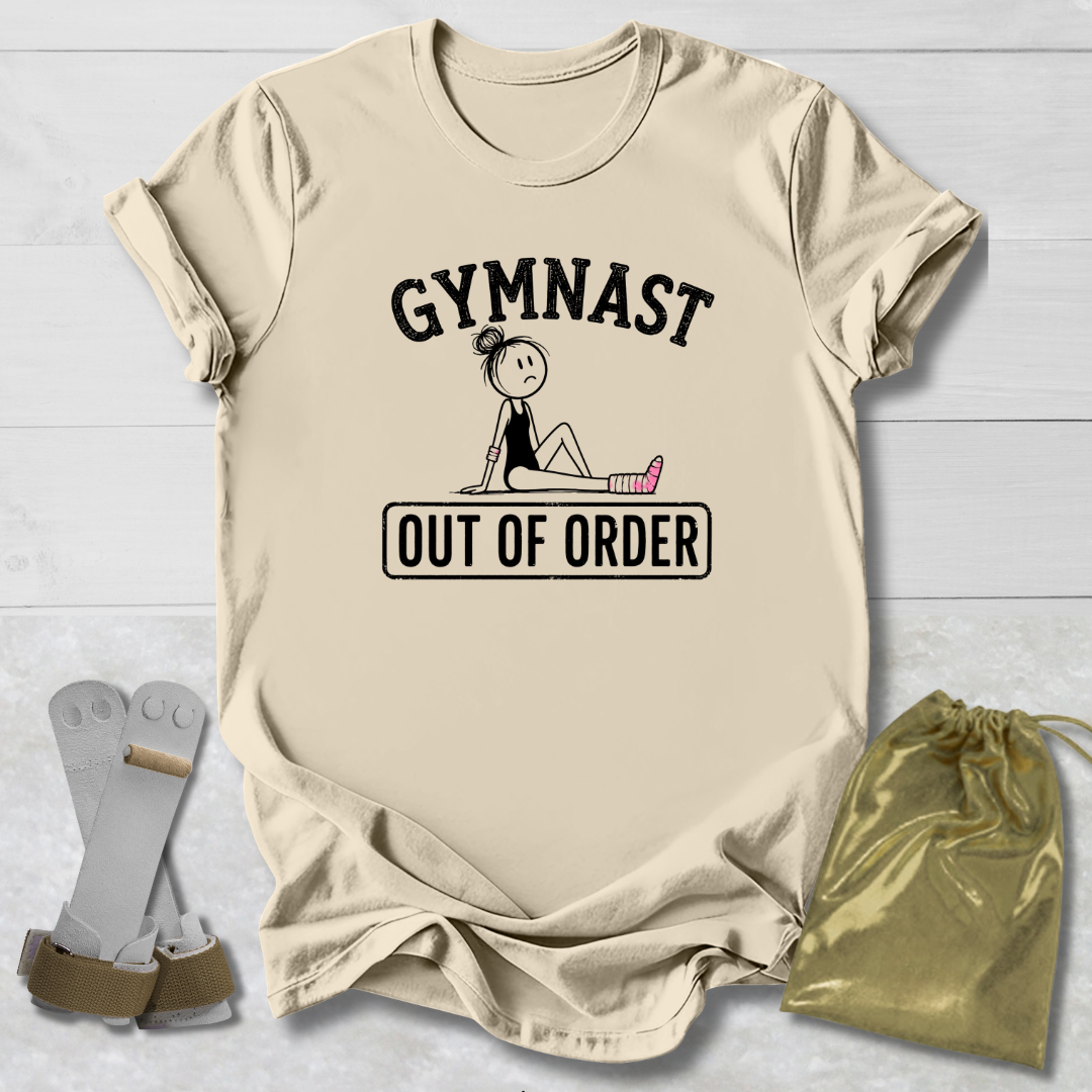 A beige graphic t-shirt laid flat on a white wood background, featuring the text 'GYMNAST OUT OF ORDER' with a simple gymnast illustration. A pair of gymnast straps and a gold drawstring bag are placed beside the shirt as styling props.